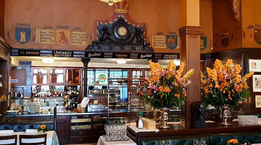 Un classico bar in stile europeo, con boiserie ornate, un grande orologio, stemmi decorativi, scaffali a specchio pieni di bottiglie e vasi di fiori arancioni vivaci sul bancone e sui tavoli.
