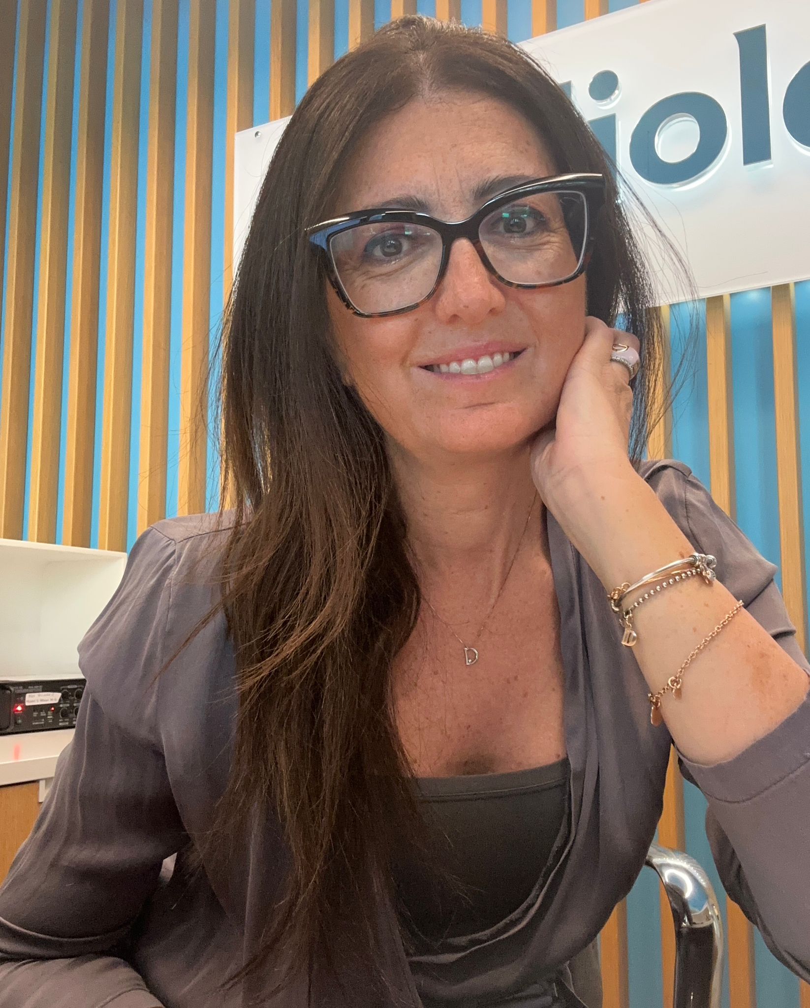 Una donna con lunghi capelli castani e occhiali neri sorride alla telecamera, appoggiando il mento sulla mano. Indossa un top grigio, braccialetti d'oro ed è seduta davanti a una parete a righe blu e legno.