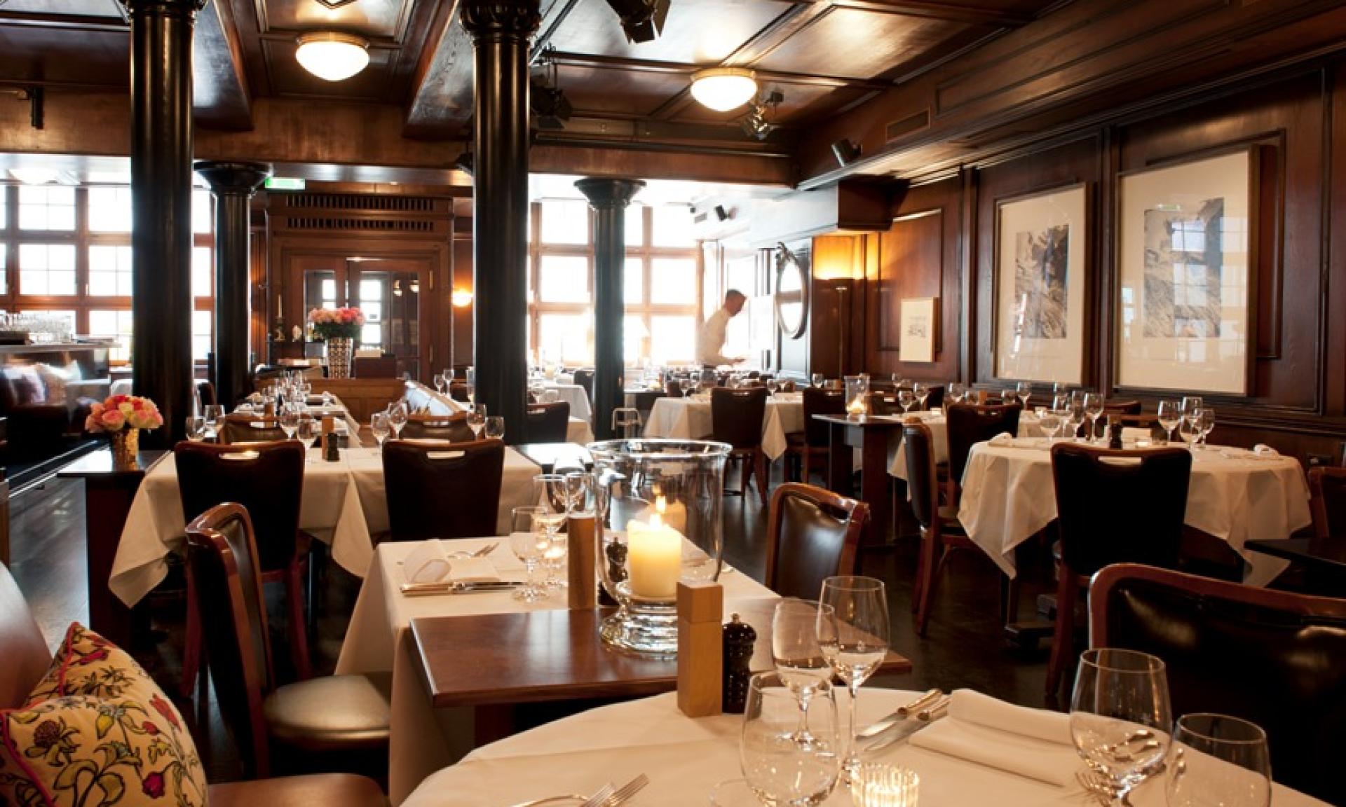 Interno di un ristorante elegante con pannelli in legno scuro, tovaglie bianche, tavoli apparecchiati con bicchieri e argenteria, luci soffuse e ampie finestre che lasciano entrare la luce naturale. Un membro del personale è visibile sullo sfondo.