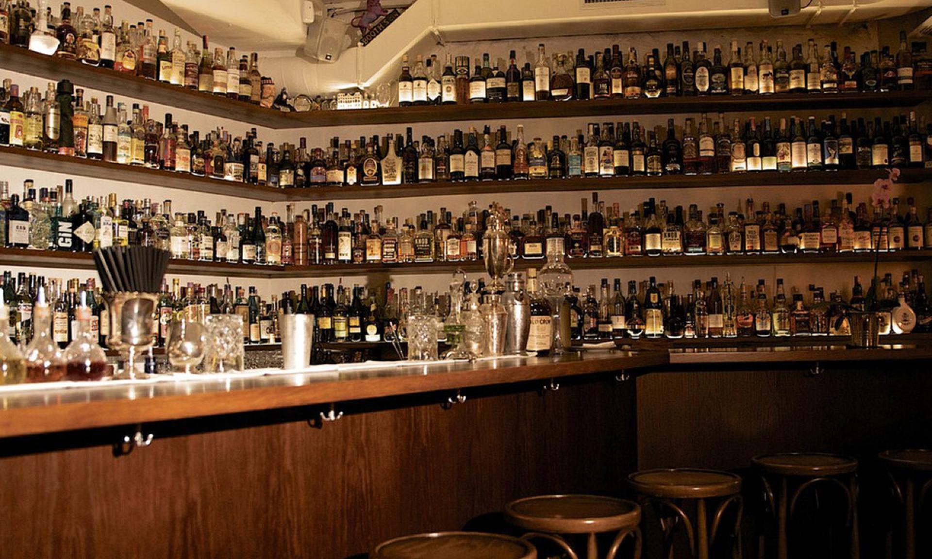 Un bar accogliente con scaffali allineati dal pavimento al soffitto con varie bottiglie di liquore. Il bancone del bar ha bicchieri, shaker e sgabelli davanti, creando un'atmosfera calda e invitante.