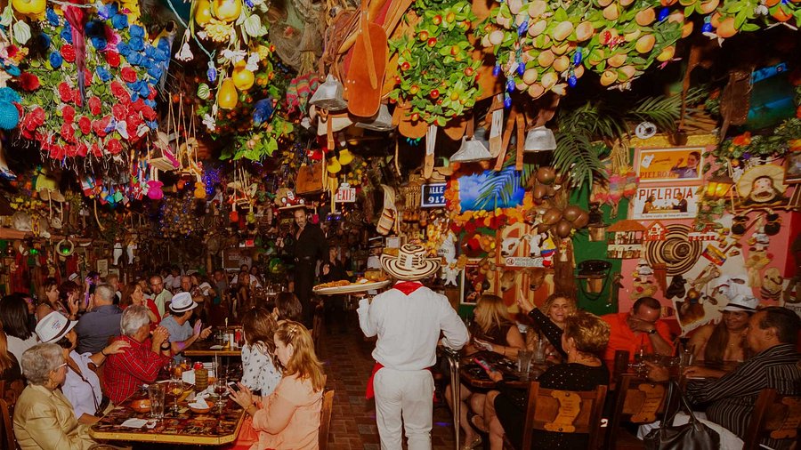 Un ristorante vivace e colorato, pieno di gente che pranza. Il soffitto e le pareti sono decorati con fiori, frutta, cappelli e ornamenti vari. Un cameriere in abiti tradizionali porta un vassoio tra la folla indaffarata e festosa.