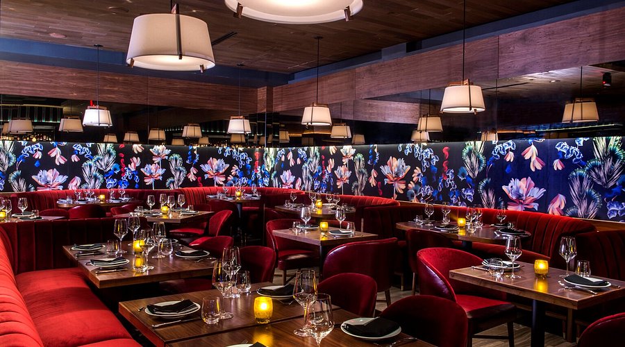 Un ristorante elegante, con cabine di velluto rosso, tavoli di legno scuro con bicchieri di vetro e una calda illuminazione a candela. I divisori con motivi floreali e le eleganti luci a sospensione creano un'atmosfera accogliente e raffinata.