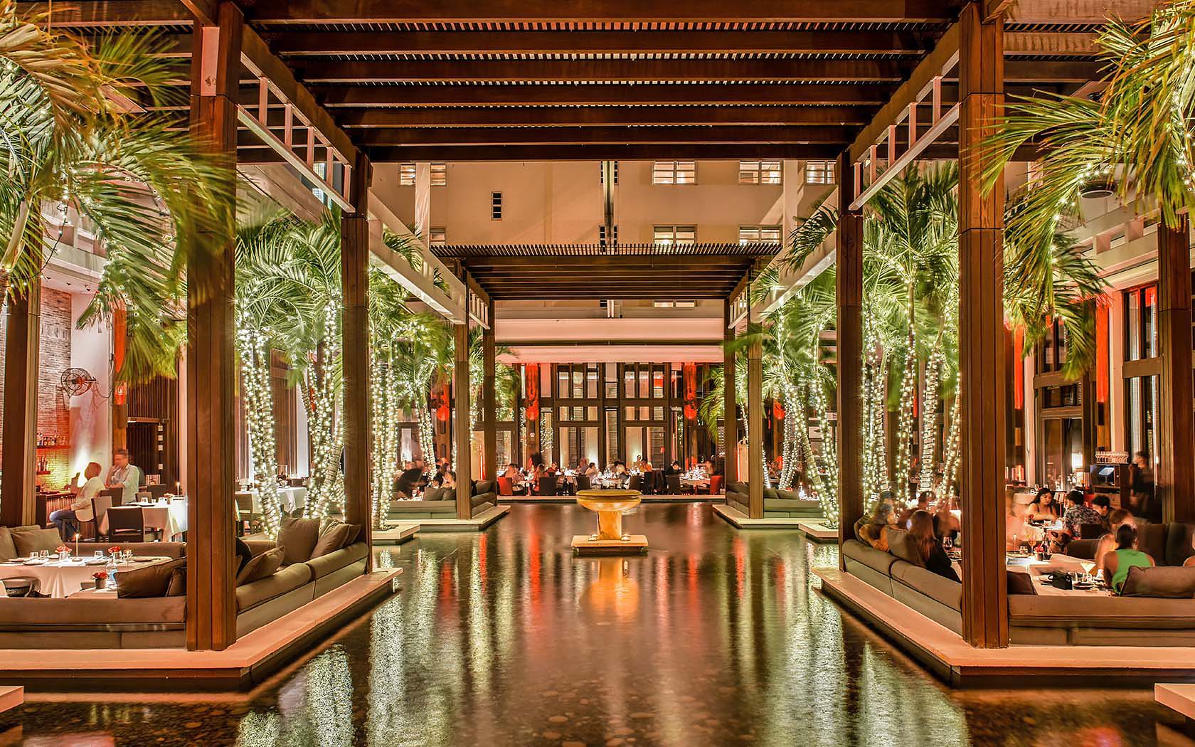 Una lussuosa hall d'albergo all'aperto con alte palme, un'illuminazione calda, luci a corda avvolte intorno alle colonne, una fontana centrale, l'acqua che riflette le luci e persone che cenano in un'atmosfera elegante e invitante.