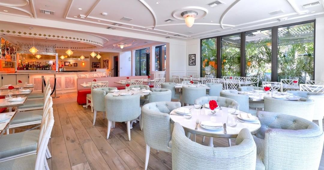 Interno di un ristorante luminoso ed elegante, con tavoli rotondi, sedie imbottite di colore chiaro, fiori rossi come centrotavola e ampie finestre che si affacciano su un verde fogliame. Sullo sfondo è visibile un bar ben illuminato.