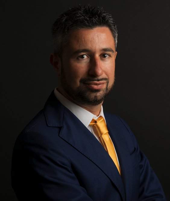 Un uomo con i capelli corti e scuri e la barba indossa un abito blu, una camicia bianca e una cravatta gialla. Posa su uno sfondo scuro con le braccia incrociate e un leggero sorriso.