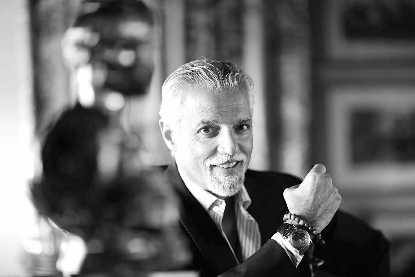 Un uomo sorridente, con capelli grigi e barba, siede in casa, indossando un abito e un orologio. Alza il braccio sinistro e in primo piano è visibile una statua sfocata. La foto è in bianco e nero.