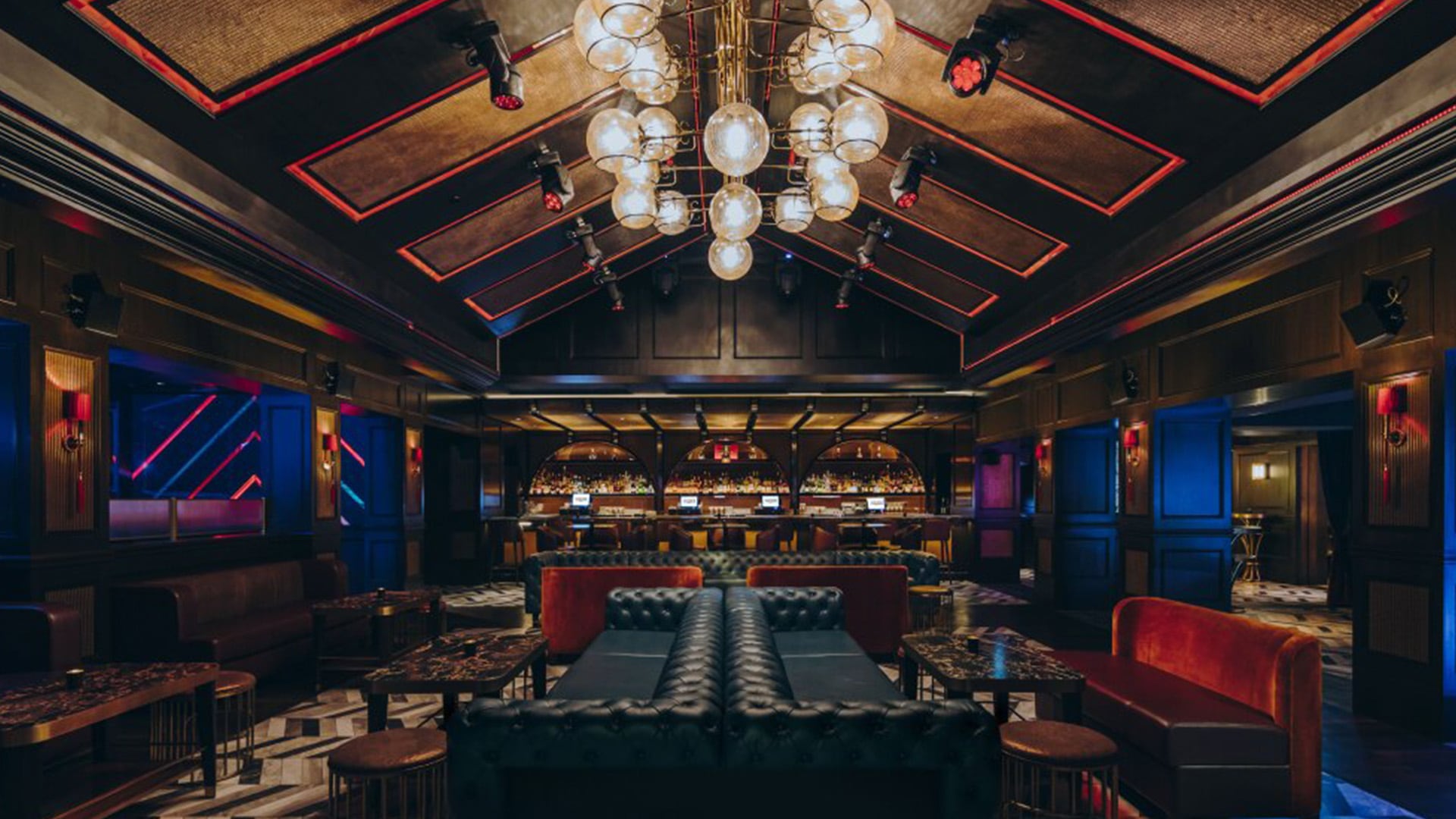 Un bar lounge elegante e poco illuminato con divani in pelle scura, sedute in velluto rosso, un lampadario geometrico e un lungo bancone foderato di bottiglie sullo sfondo. L'arredamento è caratterizzato da legno elegante, accenti dorati e illuminazione d'atmosfera.