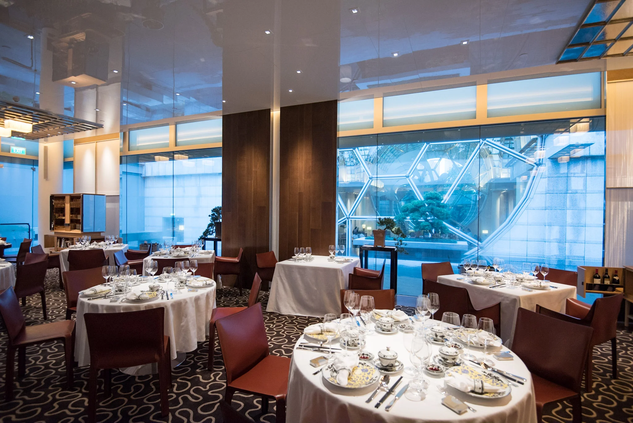 Interno di ristorante elegante con tavoli rotondi apparecchiati con tovaglie, porcellane e bicchieri bianchi. Le grandi finestre rivelano una struttura architettonica moderna e il verde esterno. L'atmosfera è luminosa e sofisticata.