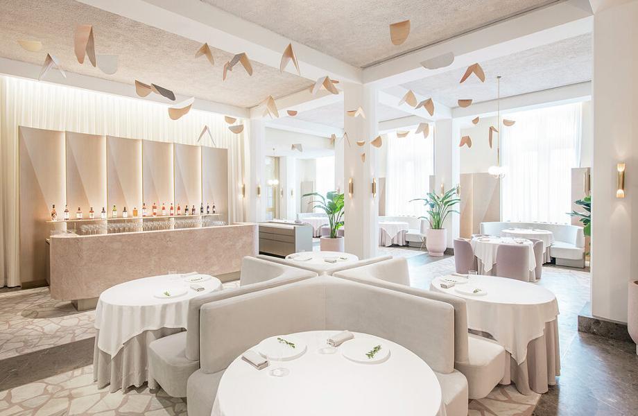Un ristorante moderno ed elegante, con tavoli rotondi ricoperti di stoffe bianche, cabine curve di colore grigio chiaro, arredamento minimale e mobili astratti appesi. Le luci soffuse e le ampie finestre creano un'atmosfera luminosa e ariosa.