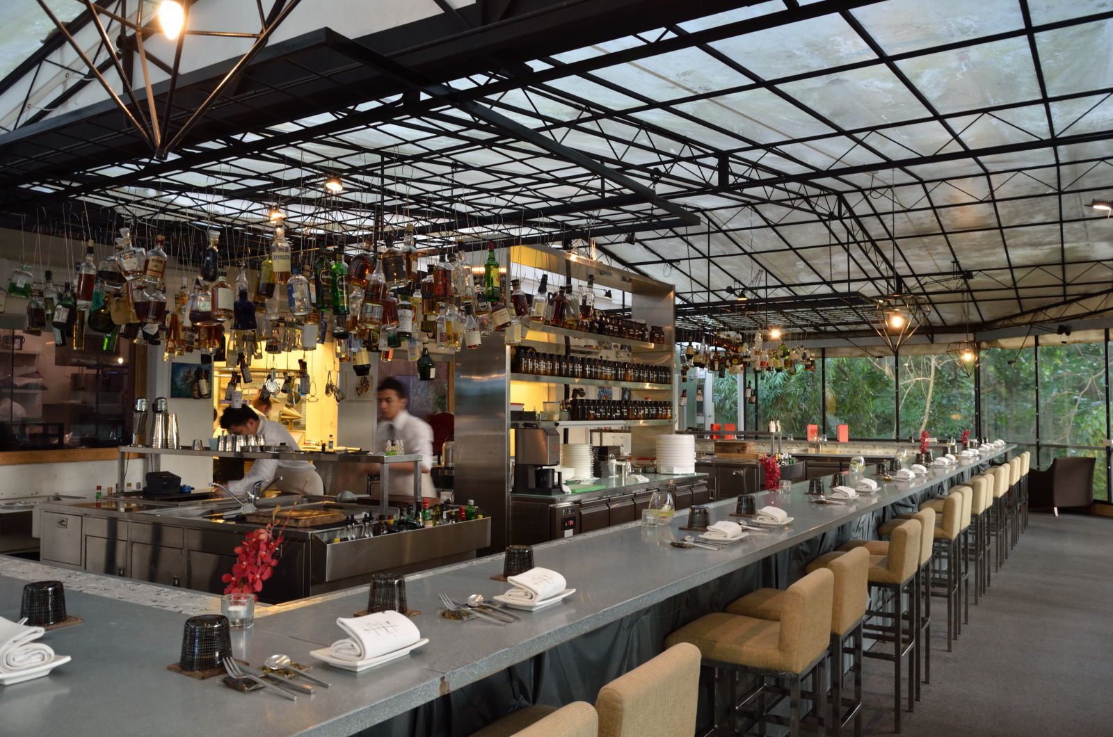 Un moderno bar ristorante all'aperto con un lungo bancone allineato con sgabelli, coperti e bicchieri appesi. Gli chef lavorano dietro il bancone e il soffitto ha una struttura metallica geometrica con luci a sospensione. All'esterno sono visibili degli alberi.