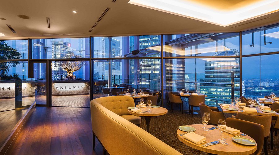 Elegante ristorante sul tetto con vetrate a tutta altezza, vista sullo skyline della città, tavoli illuminati con bicchieri e piatti e arredamento moderno con sedute accoglienti e pavimenti in legno al tramonto.