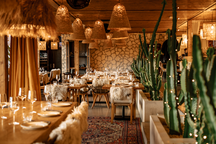 Un ristorante accogliente con tavoli in legno, sedie rivestite di pelliccia, pareti in pietra, lampade a sospensione intrecciate, grandi cactus e un'illuminazione d'ambiente calda che crea un'atmosfera invitante.