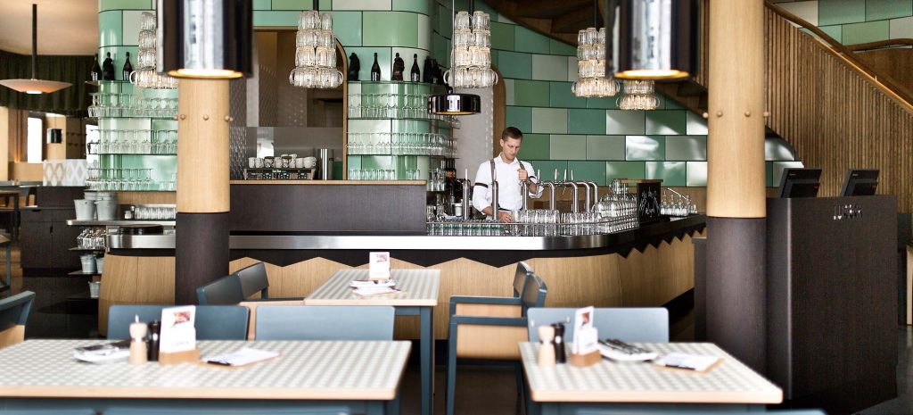Un barista si trova dietro un moderno bancone con piastrelle verdi, bicchieri e bottiglie. In primo piano ci sono diversi tavoli e sedie vuoti, con menu e condimenti su ogni tavolo. L'ambiente è luminoso ed elegante.