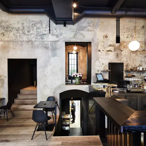 Moderna caffetteria con pareti rustiche e strutturate, mobili neri e pavimenti in legno; una scala conduce a un piano inferiore dove una persona è in piedi in una porta. Sulla destra si trova un bancone con pasticcini e attrezzature per il caffè.