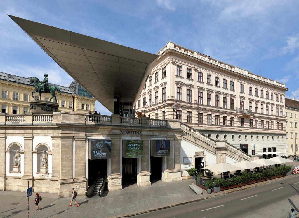 L'immagine mostra il Museo Albertina di Vienna, in Austria, con la sua moderna tettoia in vetro, l'architettura classica, la statua a cavallo, la scalinata e gli striscioni pubblicitari delle mostre. Si vedono persone vicino all'ingresso e al caffè all'aperto.