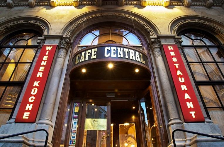 L'imponente ingresso del Café Central, con finestre ad arco, decorazioni in pietra e insegne rosse illuminate con la scritta "Wiener Küche" e "Restaurant" ai lati della porta.