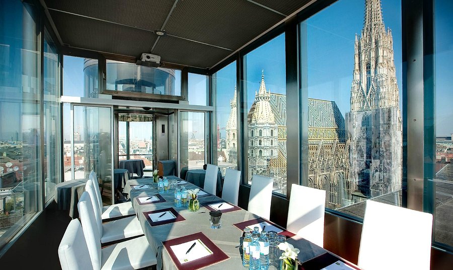 Una moderna sala conferenze con pareti di vetro, con un lungo tavolo apparecchiato per una riunione, che offre una vista panoramica di una città storica e di una grande cattedrale con torri intricate fuori dalla finestra.