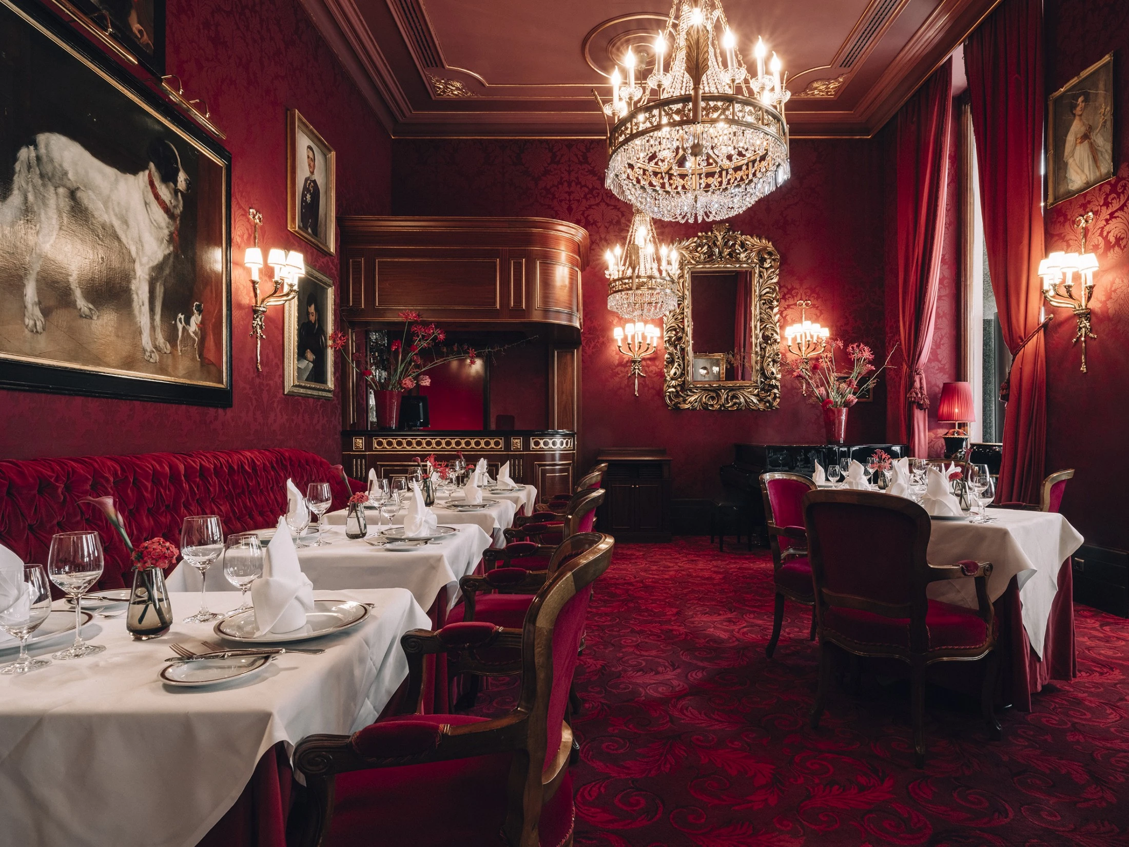 Ristorante sontuoso con pareti rosse, moquette rossa e sedie imbottite rosse. Tovaglie bianche, coperti eleganti, lampadari, specchi decorati e dipinti creano un'atmosfera lussuosa e classica.