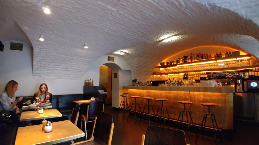 Un bar accogliente con soffitti ad arco in mattoni bianchi, un bancone illuminato d'oro con sgabelli e scaffali di bottiglie alle spalle. Due donne sono sedute a un tavolo a mangiare e parlare, mentre altri tavoli sono vuoti.