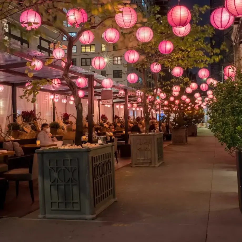 Ristorante all'aperto di sera, decorato con numerose lanterne rosa appese che creano un'atmosfera calda e invitante. Le persone cenano sotto le lanterne lungo un marciapiede cittadino fiancheggiato da alberi e piante.