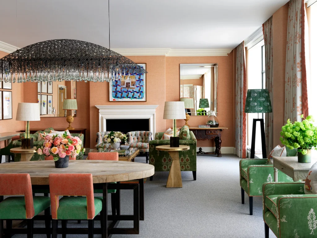 Un elegante soggiorno e sala da pranzo è caratterizzato da poltrone a fantasia verde, un tavolo in legno con sedie rosa, composizioni floreali, un caminetto bianco, ampie finestre con tende e lampade decorative sotto un moderno lampadario nero.