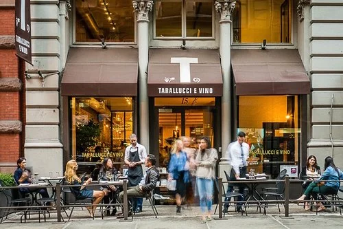 Le persone siedono e socializzano ai tavoli all'aperto davanti a Tarallucci e Vino, un ristorante italiano con grandi vetrate e tende da sole marroni in una strada cittadina. Alcune persone passano di lì, creando un'atmosfera vivace.