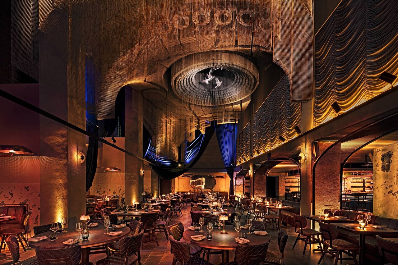 Un ristorante lussuoso e poco illuminato con arredi eleganti, tavoli rotondi apparecchiati per la cena, drappi ornati, una calda illuminazione d'ambiente e un soffitto scenografico con un lampadario circolare e decorazioni pendenti in tessuto di seta.