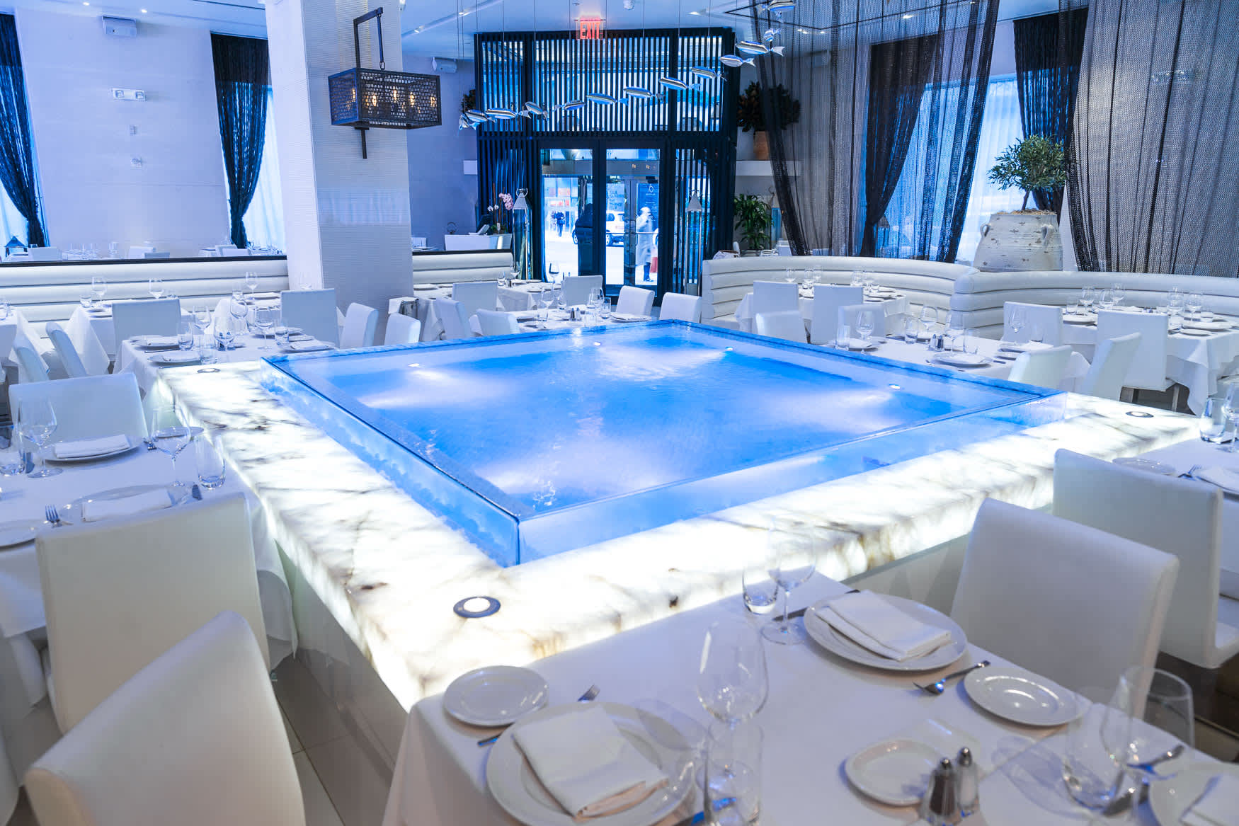 Un ristorante moderno con tavoli e sedie bianche, caratterizzato da un grande specchio d'acqua quadrato illuminato al centro, circondato da tavoli da pranzo elegantemente apparecchiati.