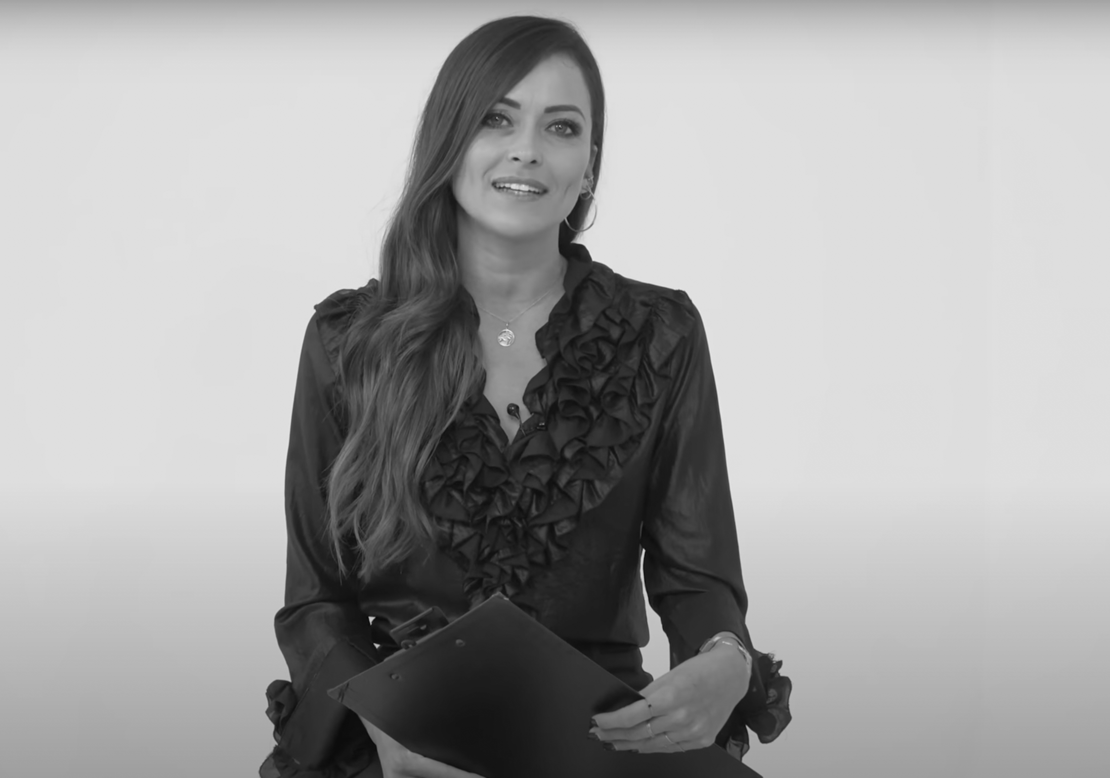 Una donna con i capelli lunghi, che indossa una camicetta arricciata, siede sorridente e tiene in mano un blocco per appunti. Lo sfondo è semplice e di colore chiaro. L'immagine è in bianco e nero.