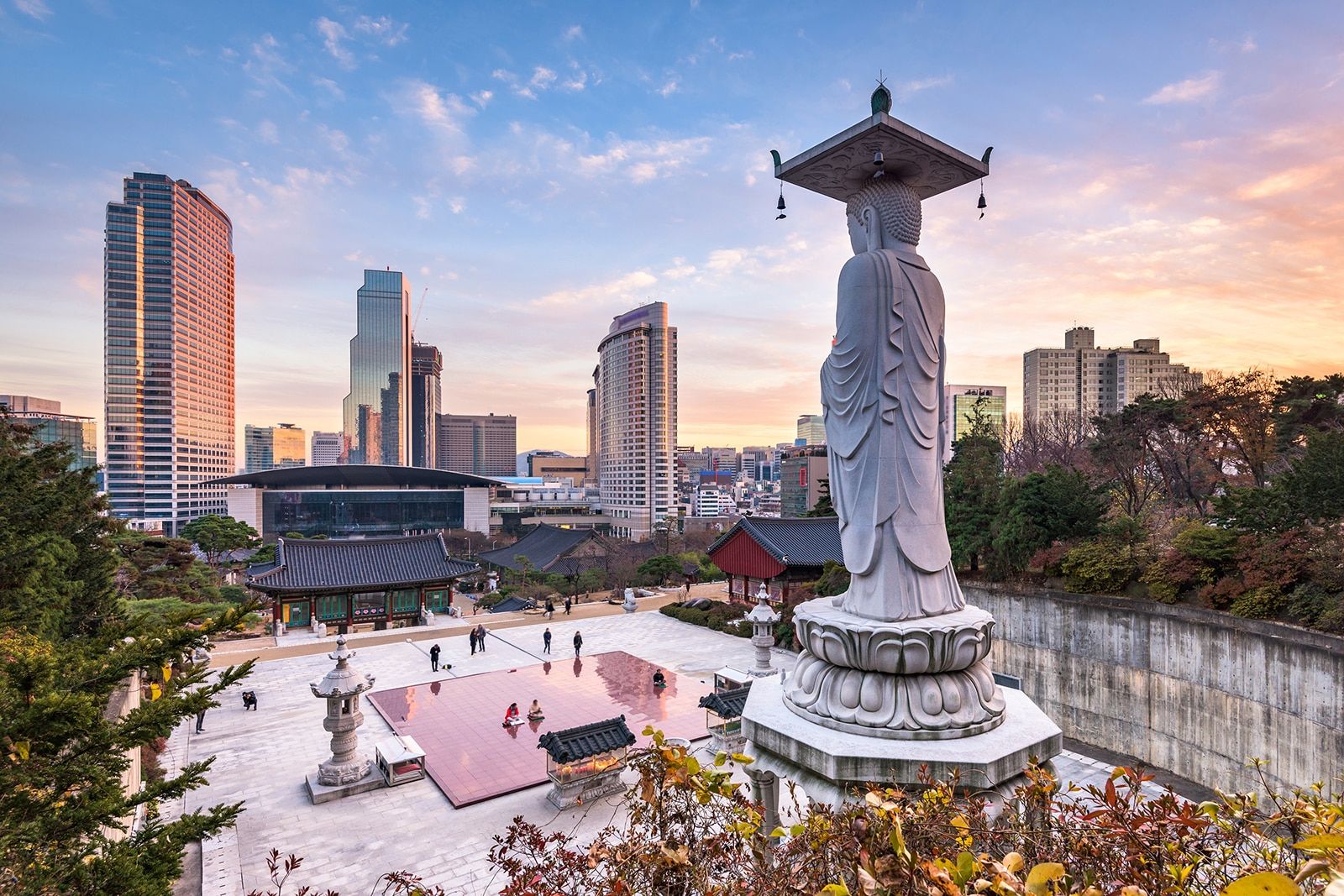 A large white Buddha statue overlooks a modern cityscape with tall buildings, a temple complex below, and people walking in the courtyard at sunset in Seoul, South Korea.
