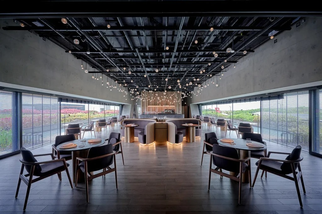 Interno di un ristorante moderno con grandi finestre su entrambi i lati, pavimenti in legno, tavoli rotondi con sedie e luci decorative sospese sul soffitto. La luce naturale riempie l'ampia ed elegante zona pranzo.
