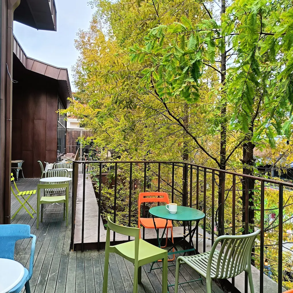 A small outdoor balcony with colorful chairs and tables overlooks lush green trees. A mug sits on one table, and the wooden floorboards and metal railing add a cozy atmosphere.