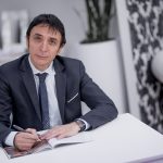 Un uomo in giacca e cravatta è seduto a una scrivania bianca, con una penna in mano e un quaderno su cui scrivere. Sullo sfondo ci sono vasi decorativi, una pianta e una parete decorata.