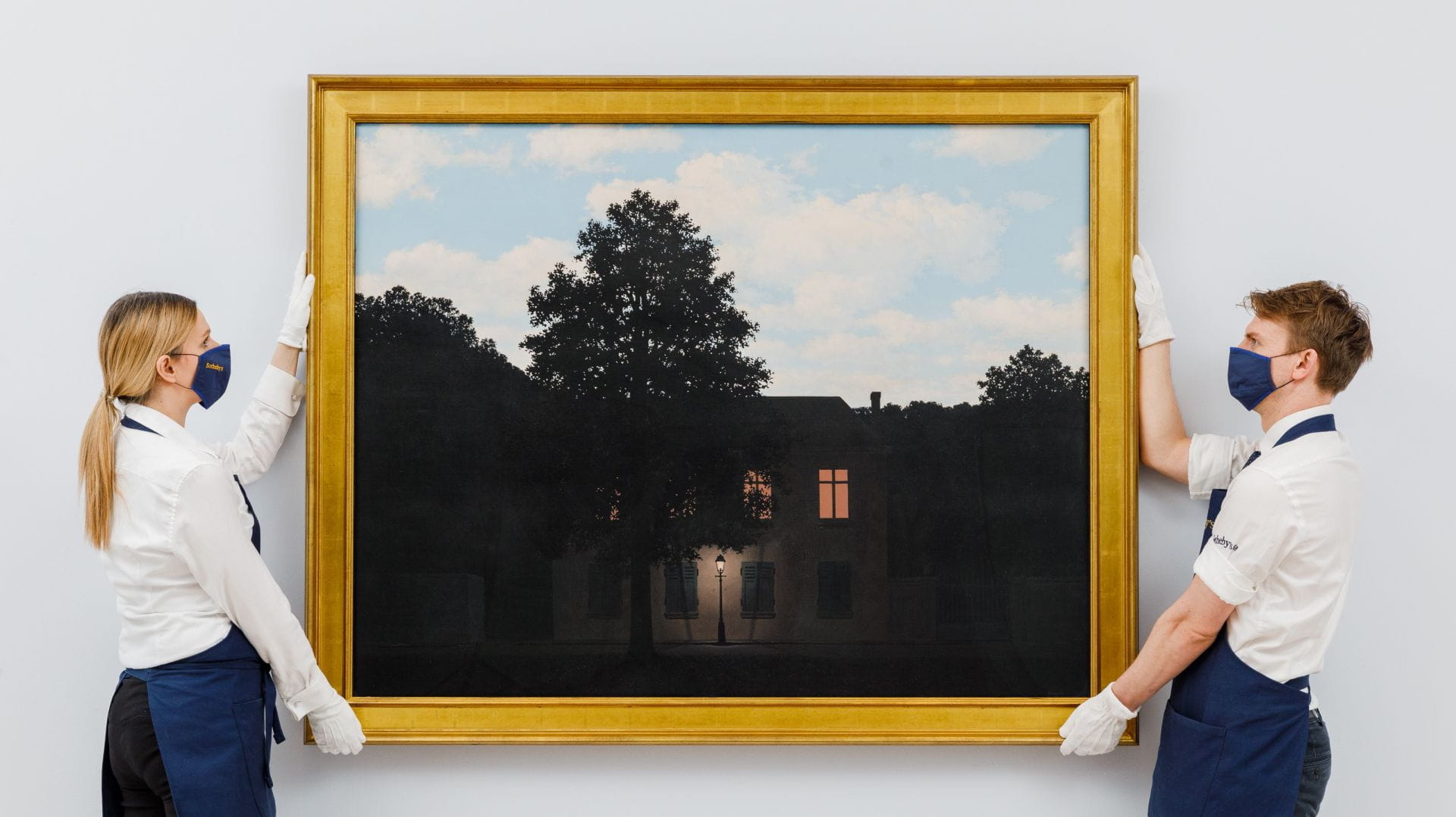 Due individui che indossano maschere e guanti tengono in mano un grande dipinto incorniciato che ricorda Magritte, raffigurante una casa al tramonto nascosta dagli alberi, con una finestra illuminata. Come curatori di un raro disco, sistemano delicatamente l'opera d'arte contro un muro bianco immacolato.