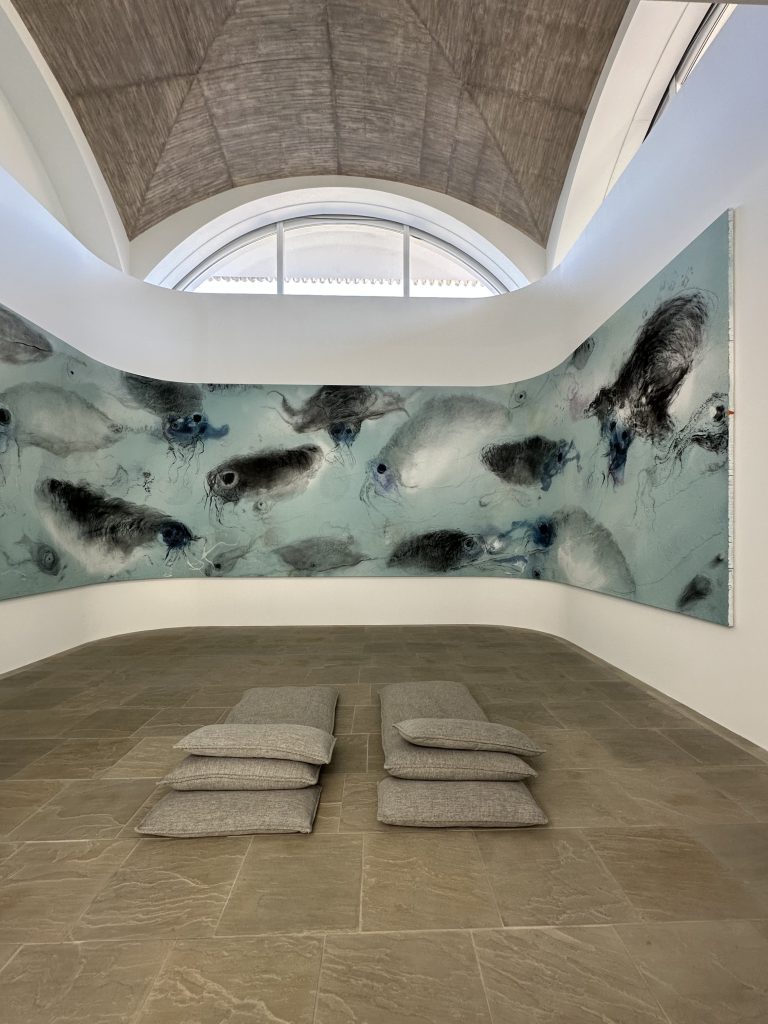 Una sala galleria minimalista presenta un grande dipinto astratto con forme fluide blu e grigie su una parete curva. Quattro cuscini grigi sono disposti su un pavimento in piastrelle di pietra, invitando alla riflessione silenziosa sotto l'alta finestra ad arco.