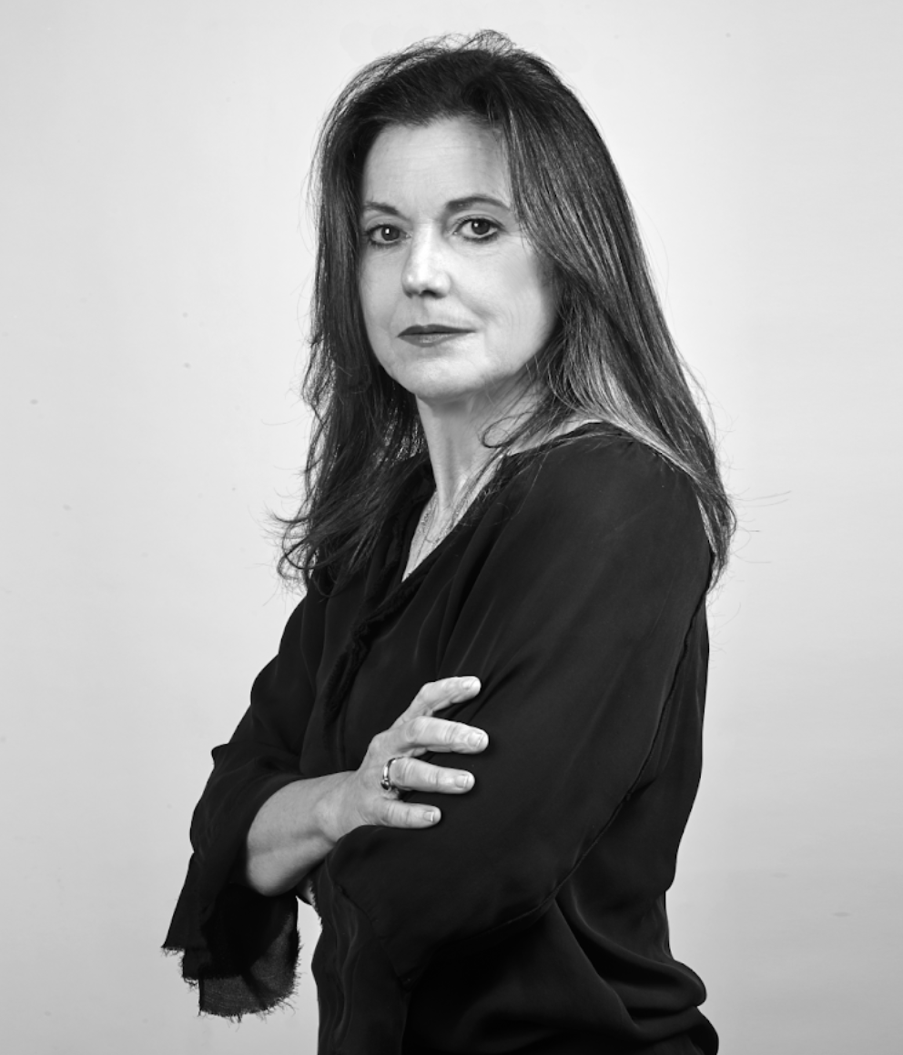 Una donna con i capelli lunghi sta in piedi con sicurezza, con le braccia incrociate, guardando la telecamera. Indossa una camicetta scura. Lo sfondo è semplice, evidenziando la sua espressione seria. L'immagine è in bianco e nero.