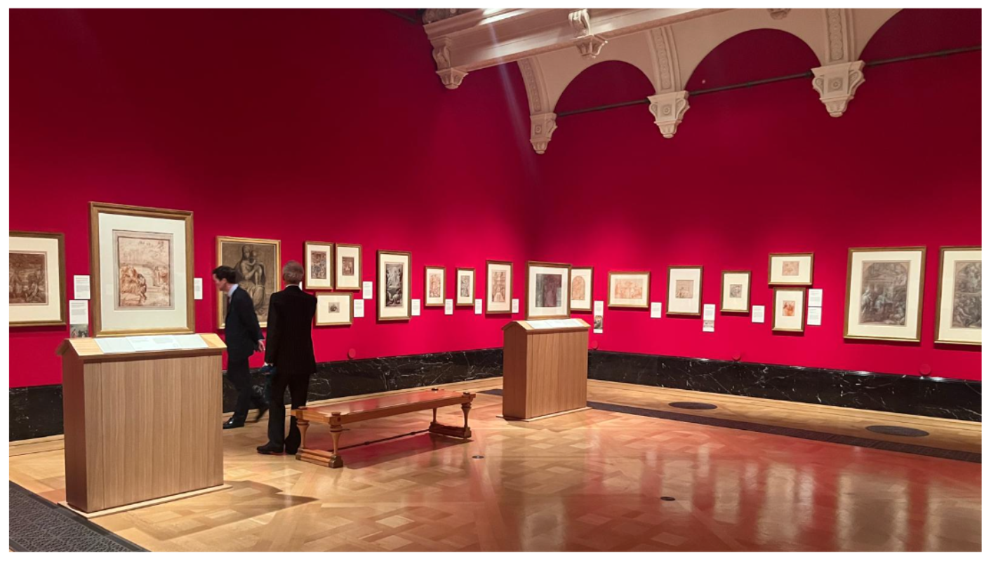 Una sala della galleria con pareti rosso acceso espone numerose opere d'arte incorniciate. Due persone stanno osservando le mostre. La sala è dotata di panche e vetrine in legno, con eleganti dettagli del soffitto ad arco.