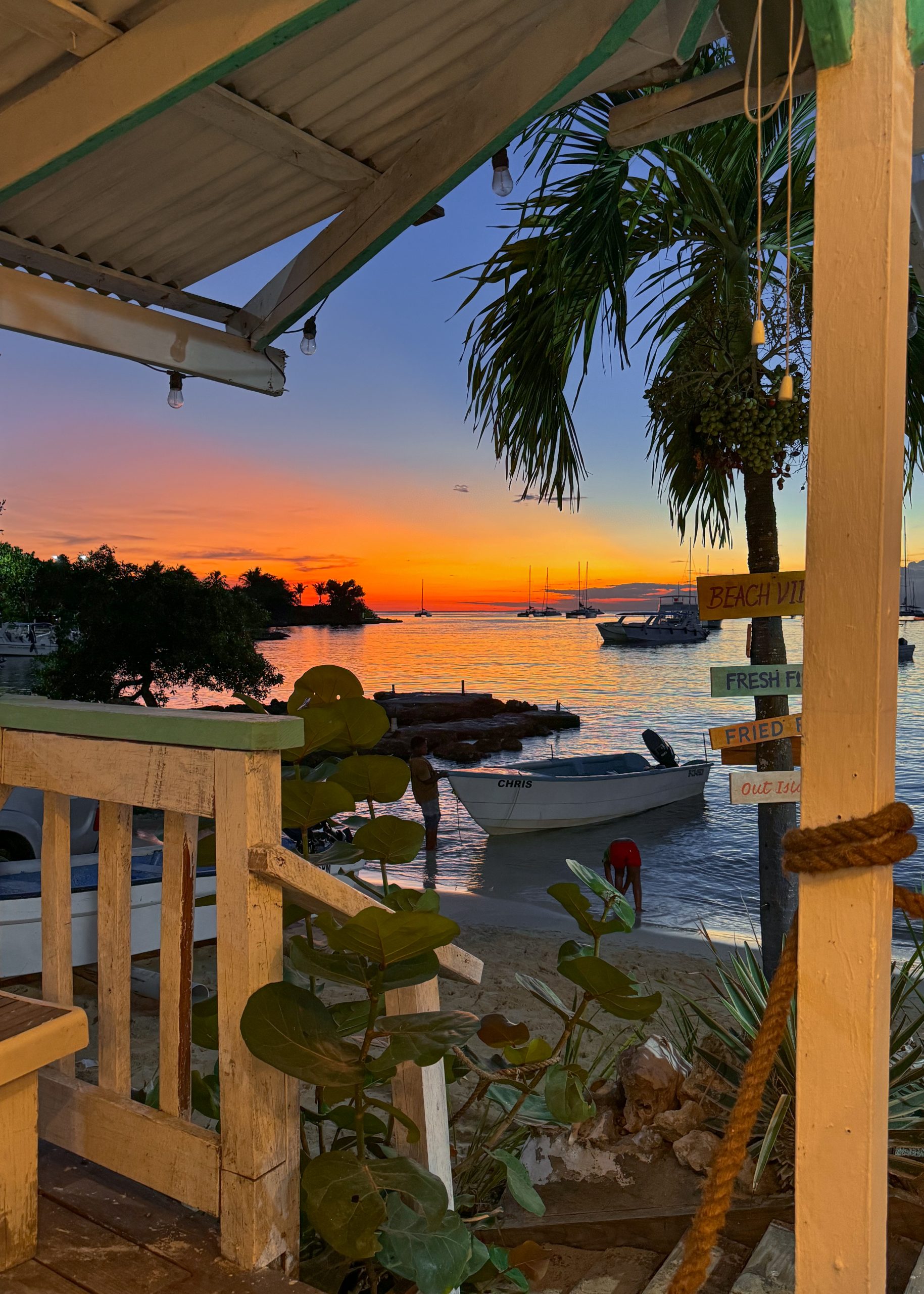 Una scena di spiaggia serena al tramonto, con un ponte di legno e una ringhiera in primo piano. L'acqua calma riflette il cielo arancione, rosa e viola. Le barche sono ormeggiate nelle vicinanze, con una palma e una vegetazione lussureggiante che si aggiungono all'atmosfera tropicale.