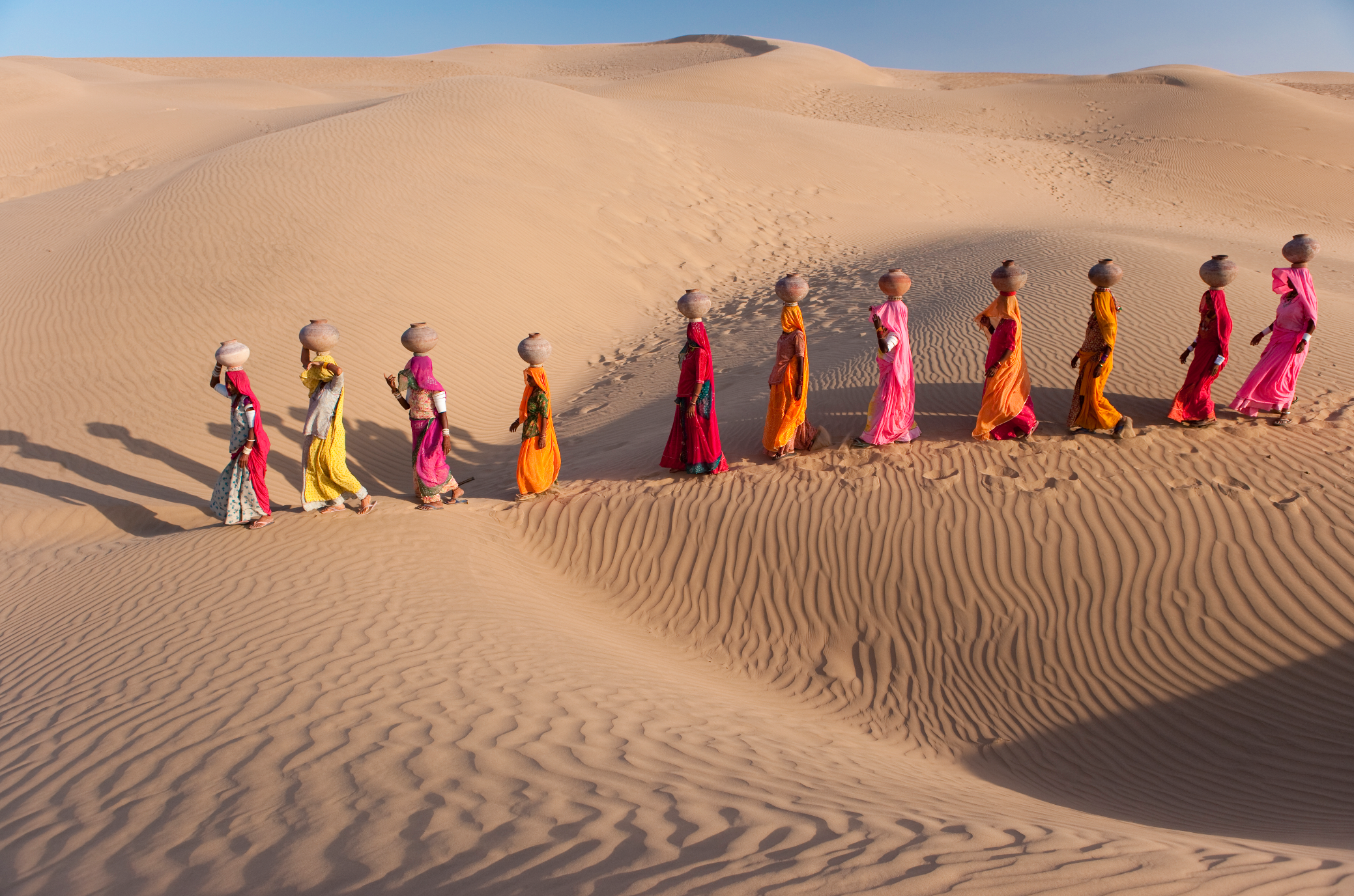 Un gruppo di donne in vivaci abiti tradizionali e foulard camminano in fila indiana sulle dune sabbiose sotto un cielo azzurro e terso. I colori dei loro indumenti contrastano con la sabbia beige, creando un effetto visivo sorprendente.