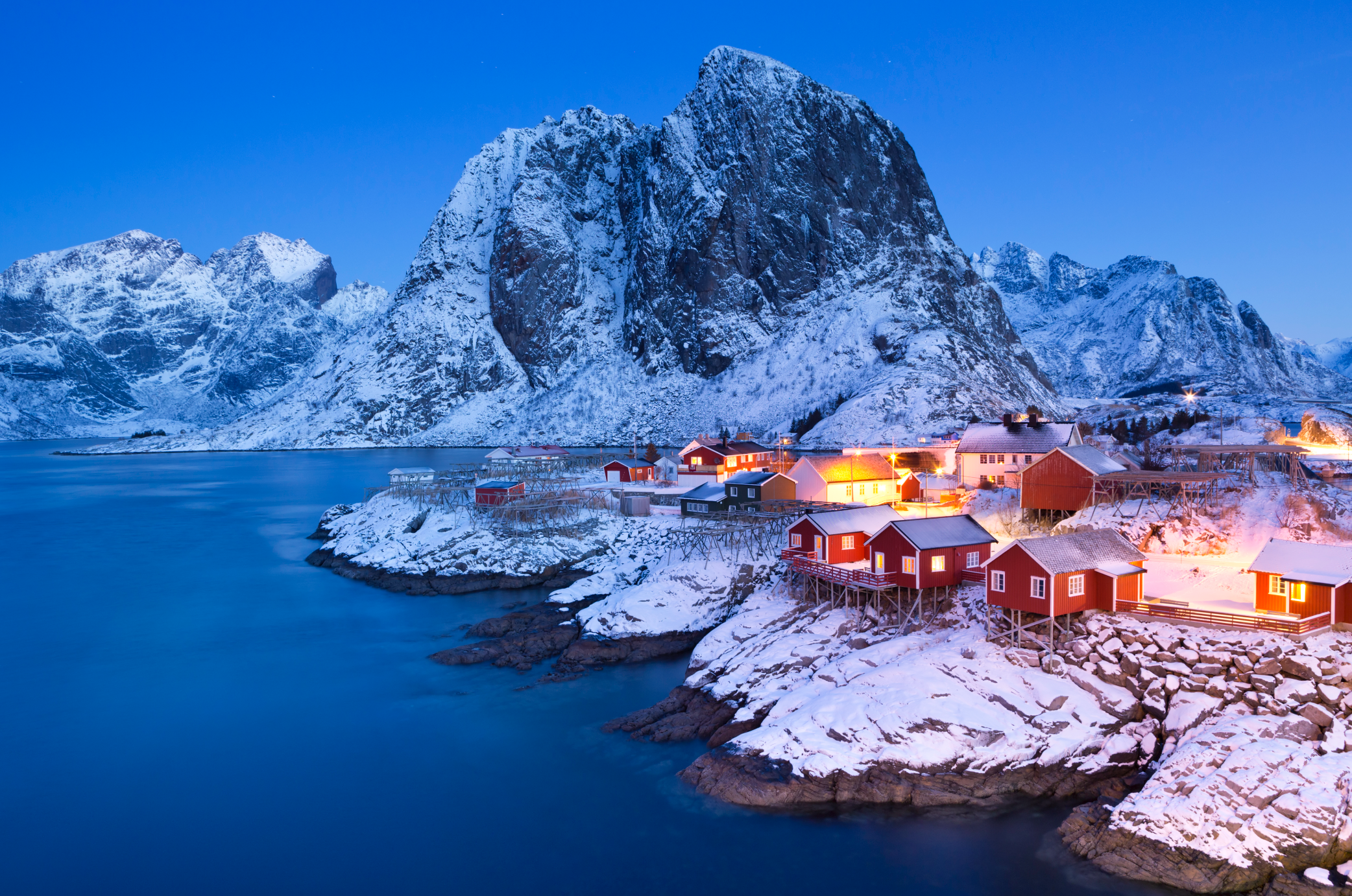 Villaggio innevato con cottage rossi lungo una costa rocciosa sotto un cielo crepuscolare. Montagne maestose si ergono sullo sfondo e luci calde brillano dolcemente dalle case, riflettendosi sull'acqua calma e azzurra.