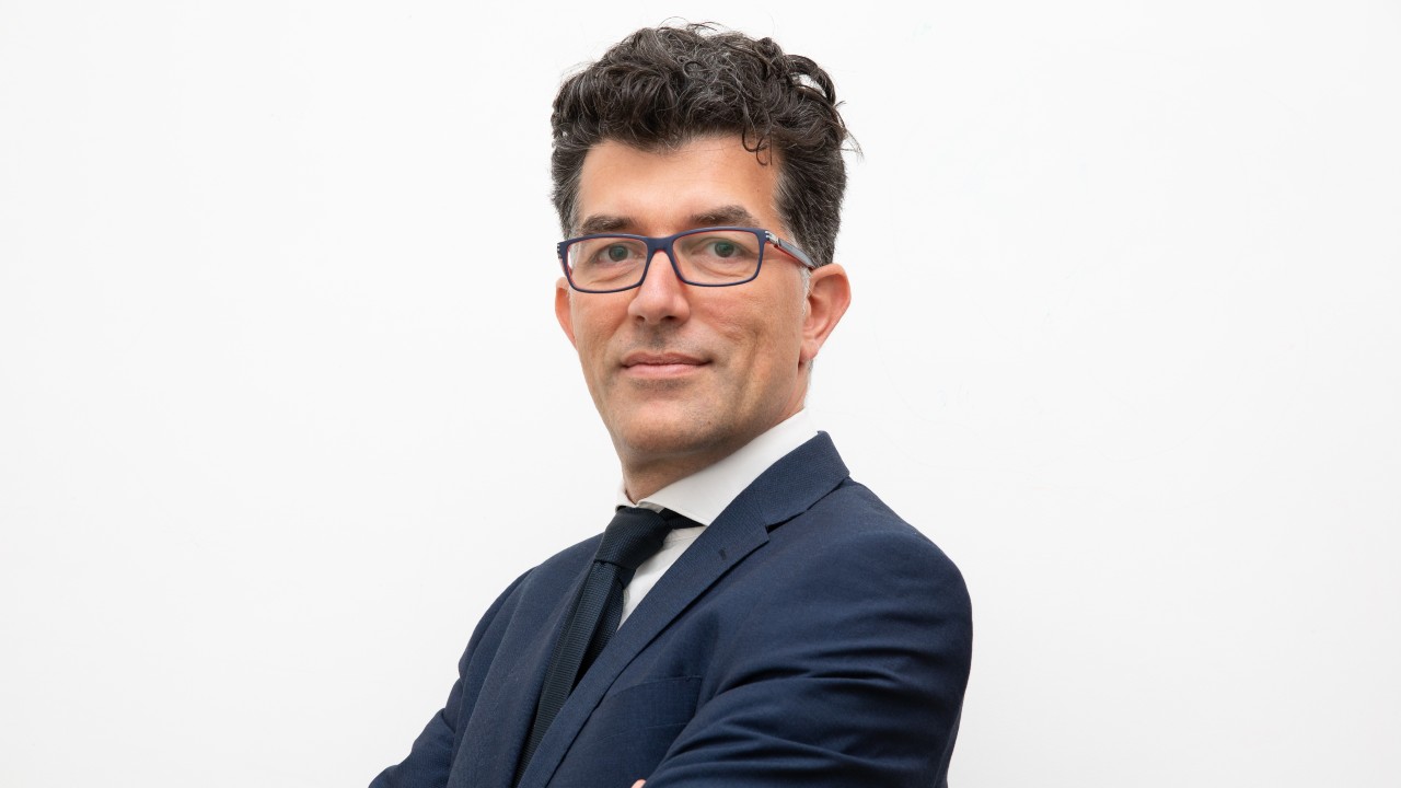 Un uomo in completo blu navy, camicia bianca e cravatta nera si erge sicuro su uno sfondo bianco semplice. Indossa occhiali e con le braccia incrociate, sorride leggermente, incarnando la professionalità spesso associata ai servizi di affitti brevi.