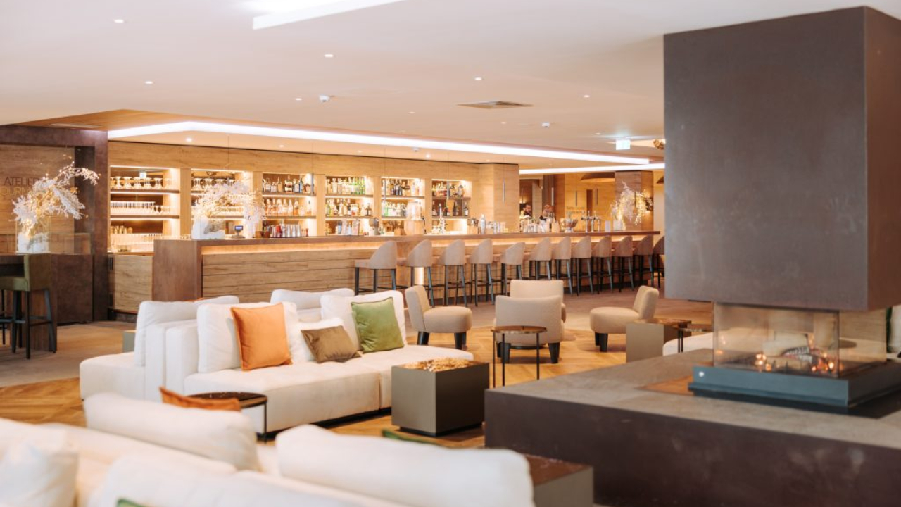 Nel cuore di Cervinia, questa moderna hall d'albergo emana un'atmosfera accogliente con divani bianchi adornati da cuscini colorati. Un camino quadrato scuro e un lungo bancone con sedie alte si trovano sullo sfondo, il tutto immerso in una calda illuminazione e circondato da piante decorative.