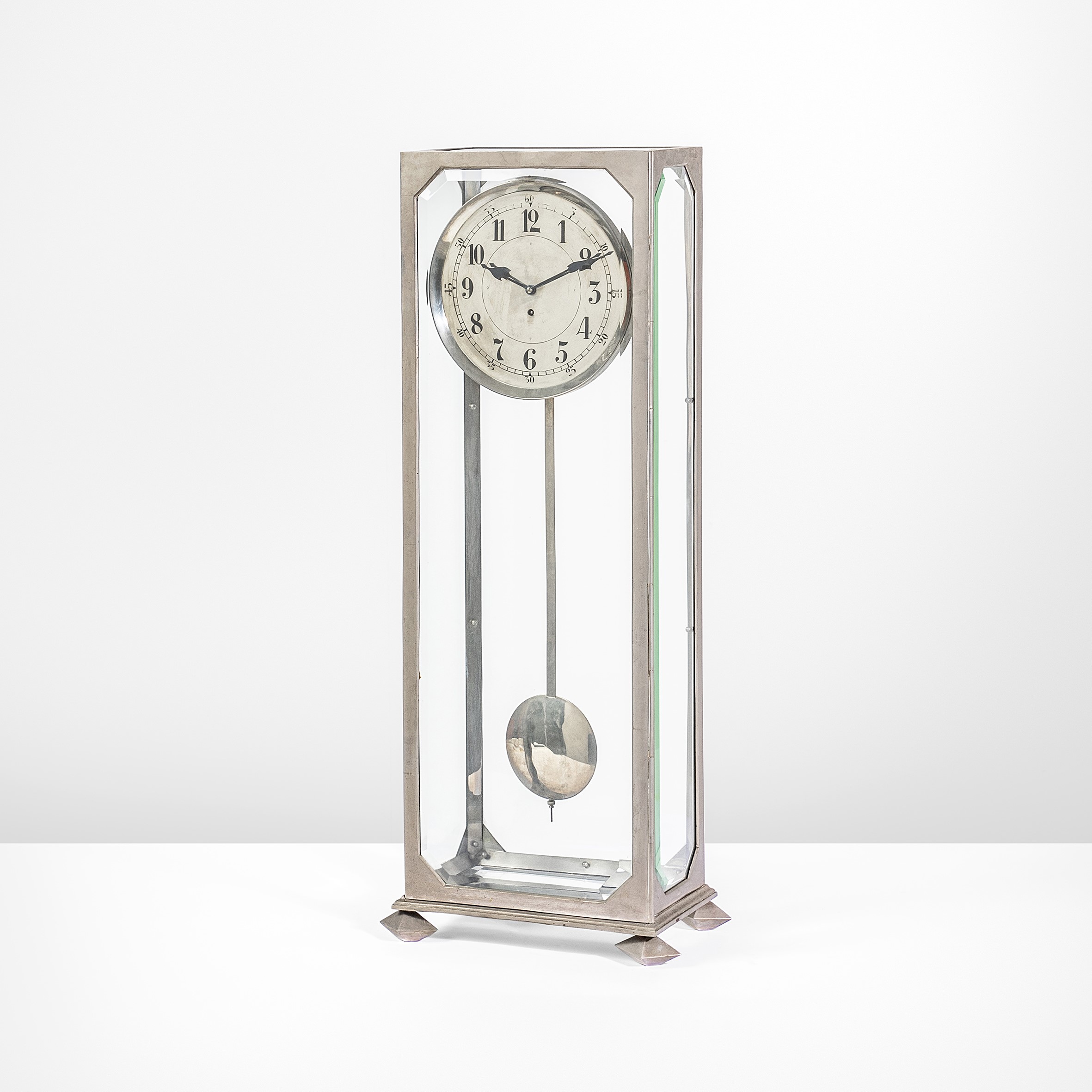 Un orologio a pendolo alto e snello in vetro e metallo con una cornice argentata si staglia su uno sfondo bianco. Il quadrante dell'orologio è rotondo con numeri neri e lancette nere ornate, mentre un pendolo lucido è appeso sotto.