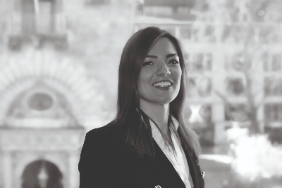 Una donna con i capelli lunghi sorride mentre indossa una giacca e una camicetta, trasudando l'eleganza di Cervinia. Lo sfondo sembra essere un paesaggio urbano sfocato in bianco e nero.