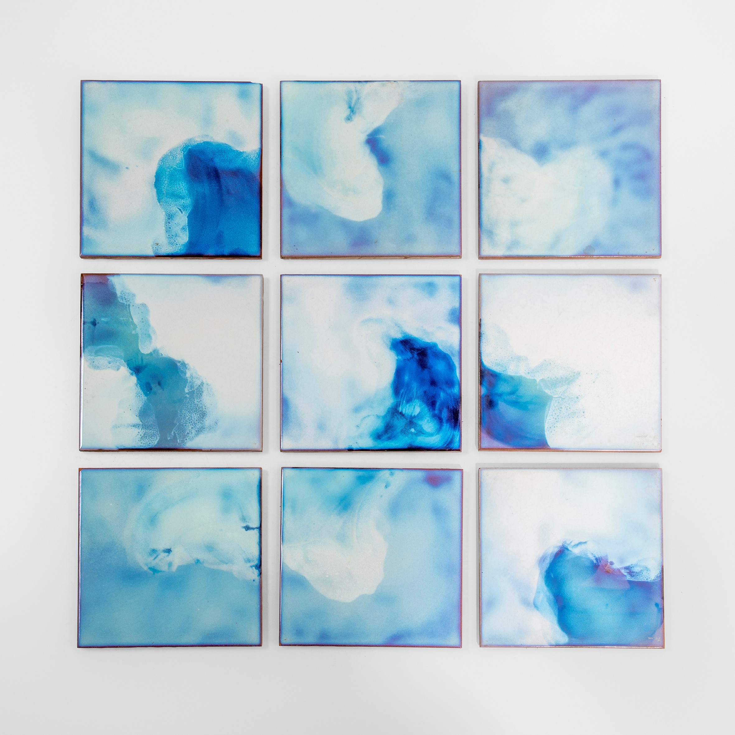 Nove piastrelle quadrate disposte in una griglia, ciascuna caratterizzata da vortici astratti di blu e bianco che ricordano le onde dell'oceano o le nuvole. Le piastrelle creano un design fluido e coeso con sfumature e motivi variabili.