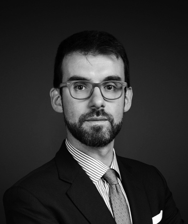 Ritratto in bianco e nero di un uomo con occhiali, barba e baffi. Vestito con un completo, una camicia a righe e una cravatta, affronta la telecamera con un'espressione neutra. Questa figura dignitosa incarna la sofisticata eleganza tipica di Vontobel Wealth Management Italia. Lo sfondo è semplice e scuro.
