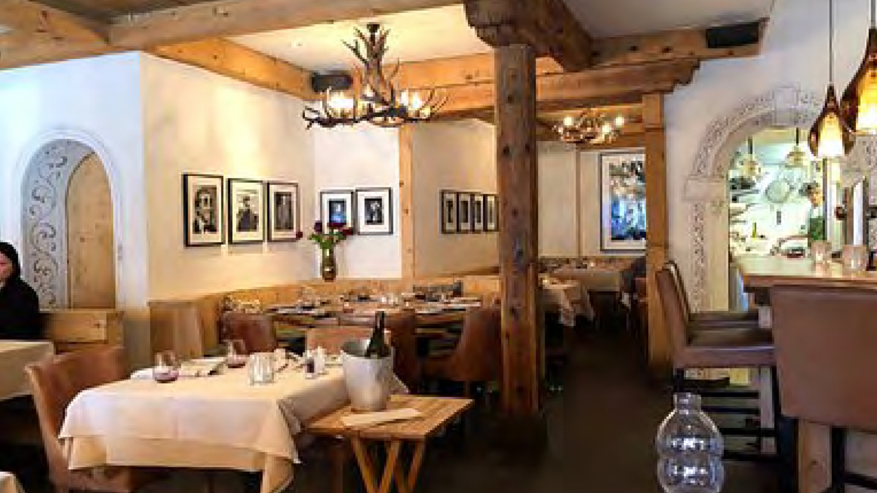 L'accogliente ristorante Sankt Moritz presenta travi in legno, lampadari di corna di cervo e foto in bianco e nero incorniciate che adornano le pareti. I tavoli sono elegantemente apparecchiati con tovaglie beige, con una bottiglia di vino fresca in un secchiello del ghiaccio su un piccolo tavolino laterale.