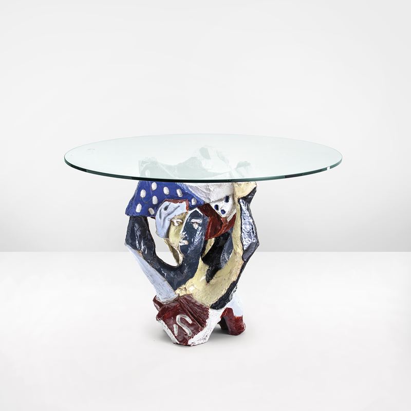 Un tavolino rotondo in vetro con una base unica e colorata. La base presenta forme astratte e scultoree in blu, rosso, giallo, nero e bianco con pois. Lo sfondo è bianco semplice, che mette in risalto il design artistico del tavolo.