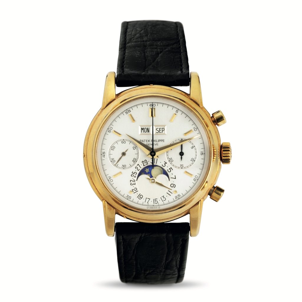 Un orologio da polso d'oro vintage con cinturino in pelle nera, che mostra giorno, data, mese e quadranti delle fasi lunari su un quadrante bianco. Ha tre quadranti secondari e due pulsanti sul lato.