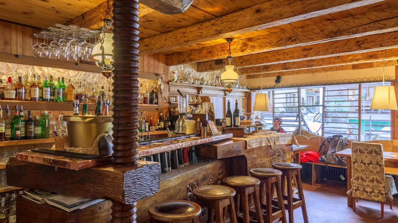 Accogliente, rustico interno di un bar a Cervinia con travi in legno e sgabelli. Scaffali pieni di varie bottiglie di alcolici. Una persona è seduta a un tavolo vicino alla finestra, dove entra la luce naturale. Atmosfera calda e invitante con lampade a sospensione.